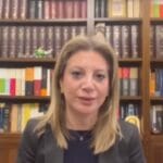 Professional woman with blond hair in a dark blazer speaks in front of a tall wooden bookshelf filled with colorful and dark books.