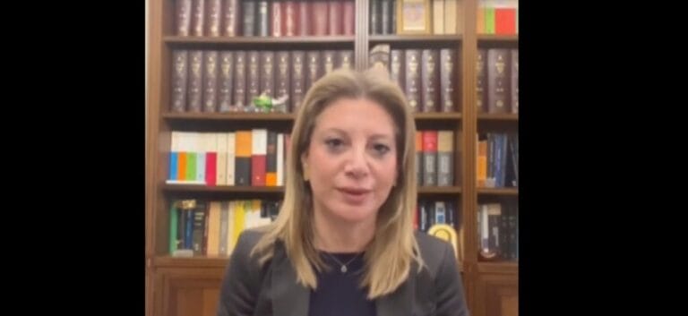 Professional woman with blond hair in a dark blazer speaks in front of a tall wooden bookshelf filled with colorful and dark books.