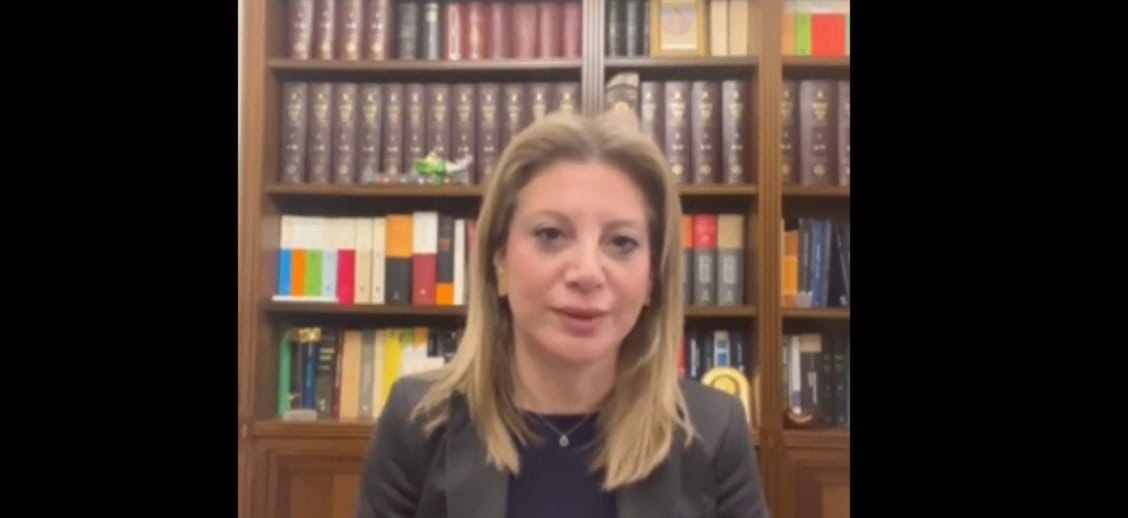 Professional woman with blond hair in a dark blazer speaks in front of a tall wooden bookshelf filled with colorful and dark books.