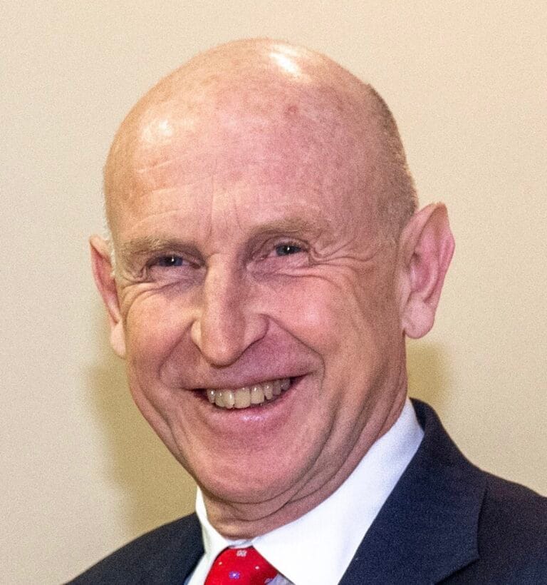 Smiling older man in a dark suit and red tie, facing the camera in a head-and-shoulders portrait.